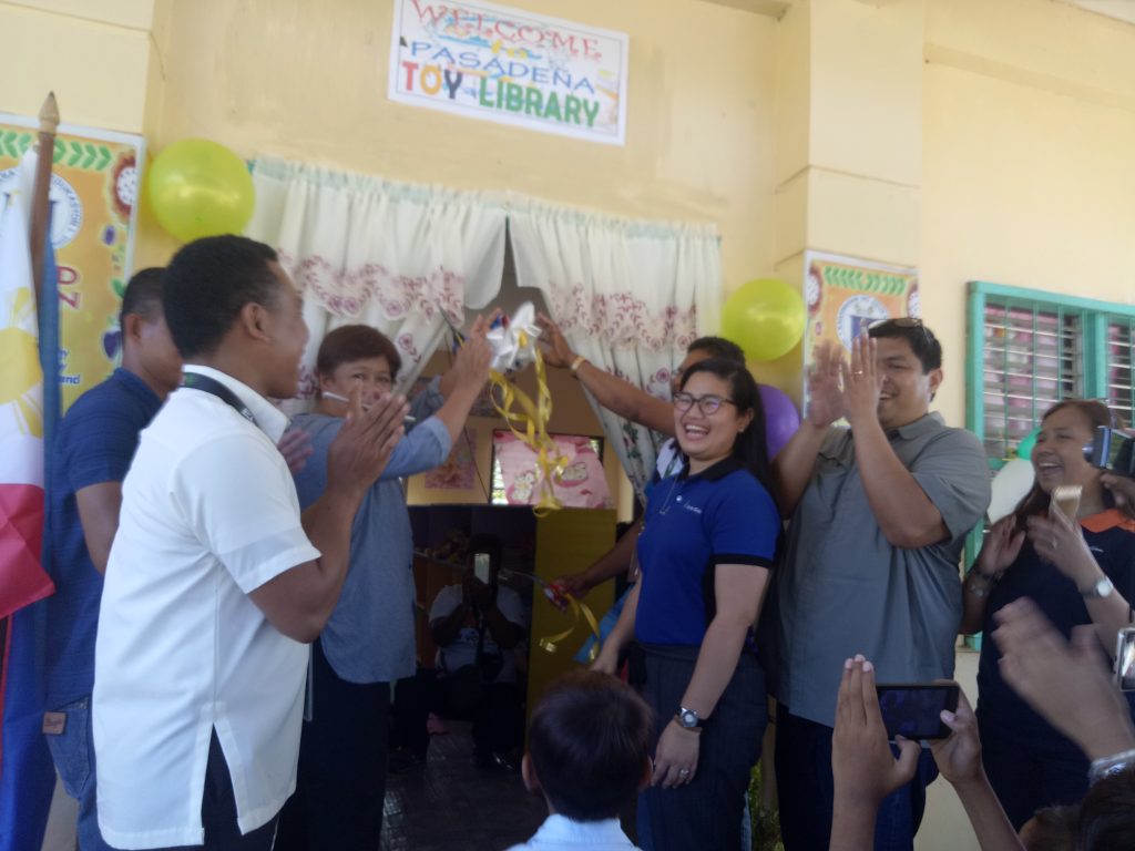 El Nido Vice Mayor Leonor Corral (third from left) and Department of Education District Supervisor Daniel Belleza (partly covered) cut the ceremonial ribbon opening the Pasadena Elementary School toy library in El Nido, Palawan as (from left, foreground) Pasadena head teacher Marlon Batoy, Lamoro Barangay Captain Dario Campuid, the Ayala Coop’s Sarah Razonable, and the Philippine Toy Library’s Edsel Ramirez and Chiara Cruz applaud.